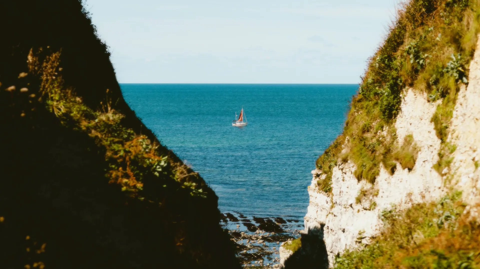 A lone sailing vessel on still water beneath dramatic coastal cliffs
