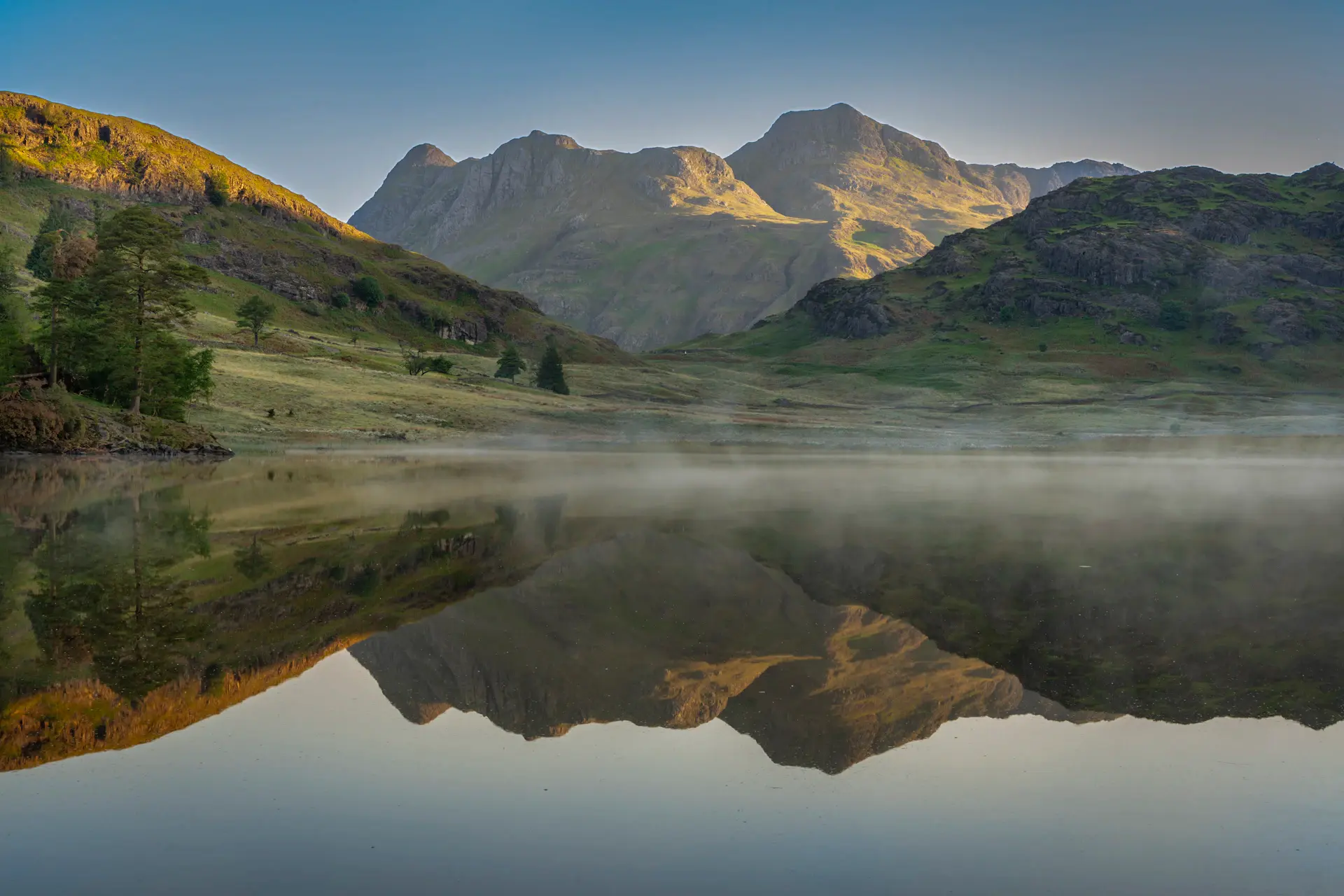 Wide view of a calm lake with mist drifting over mirrored peaks